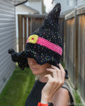A woman wearing a crocheted witch hat with a black base, pink band, and multicolored speckles, photographed outdoors in a backyard setting.