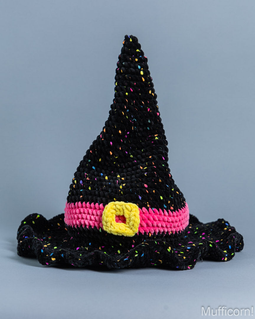 A crocheted witch hat in black yarn with multicolored sprinkle details, a bright pink band, and a yellow buckle accent, photographed against a solid gray background.