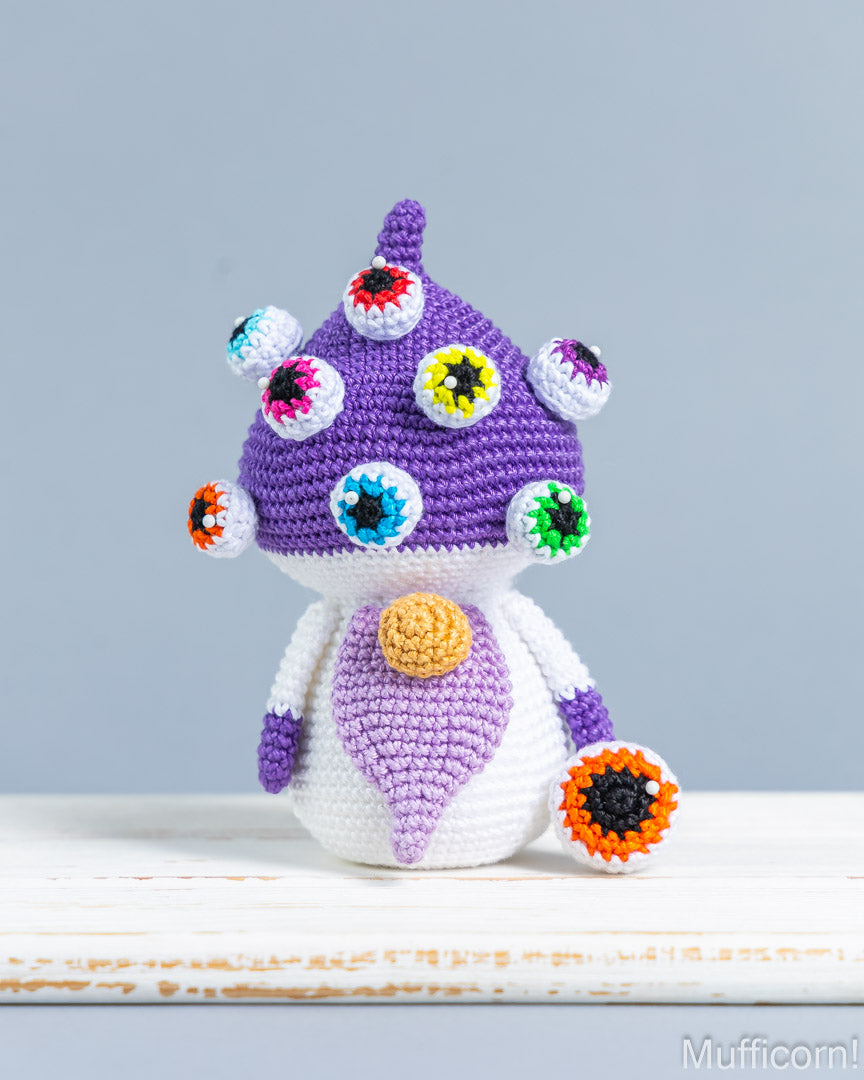 A crocheted plush toy shaped like a mushroom with a purple cap, white body, and colorful embroidered eyes and flower details, standing on a white surface against a light gray background.