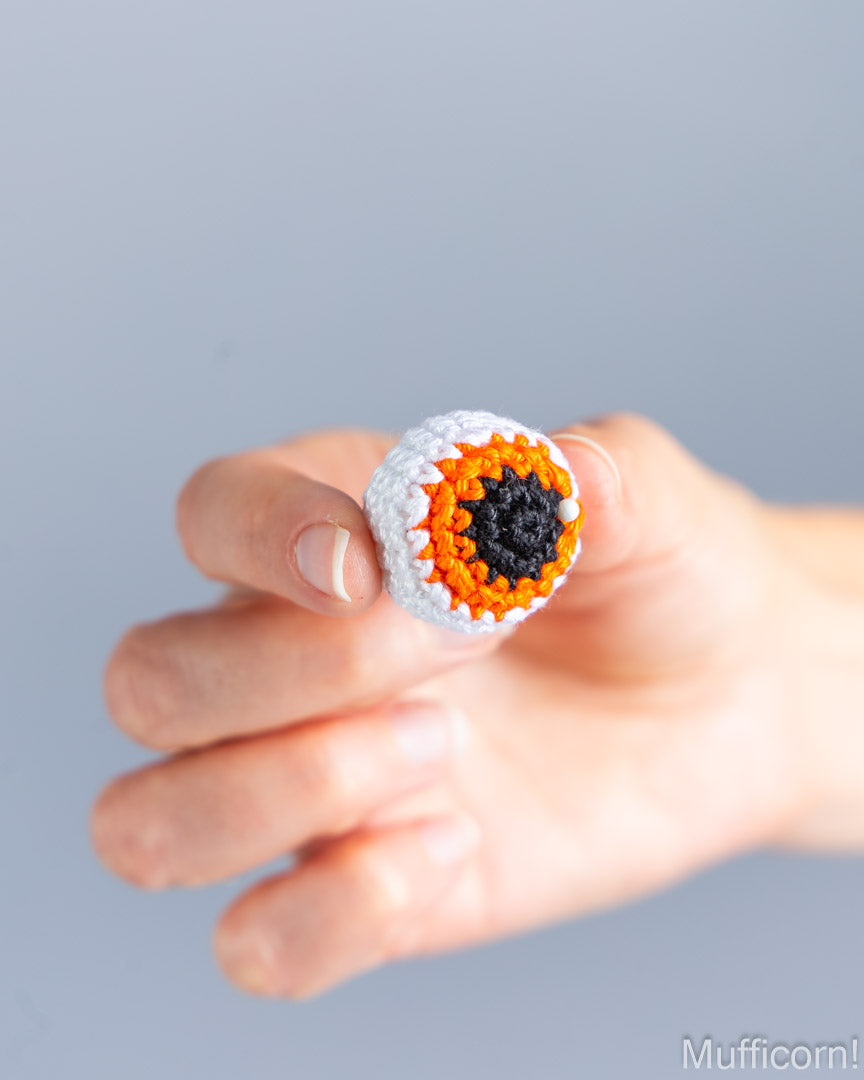 A hand holding a small crocheted ring shaped like a stylized eye, with white, orange, and black yarn, against a soft gray background.