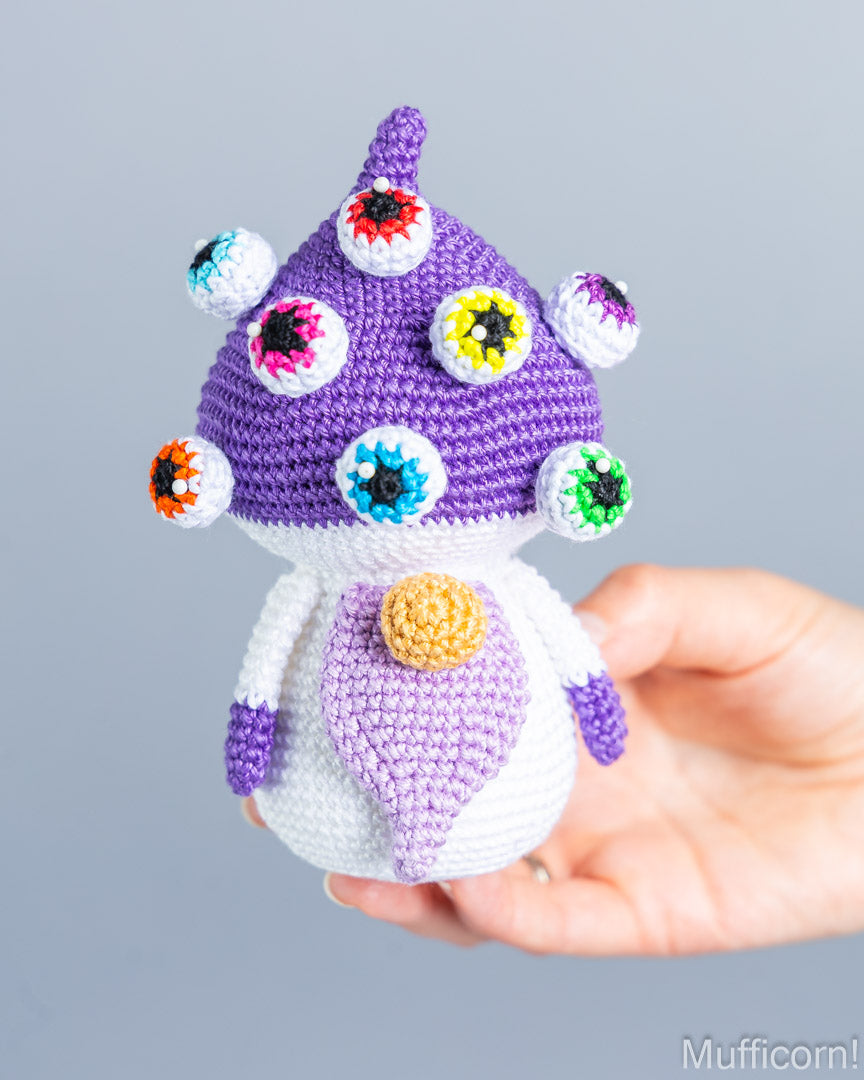 A handmade crocheted mushroom character doll with multiple colorful embroidered eyes and a yellow nose, held in a person's hand against a plain gray background.