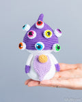 A small crocheted mushroom character doll with a purple cap, white stem, and multicolored eye details, being held in a person's hand against a plain light gray background.