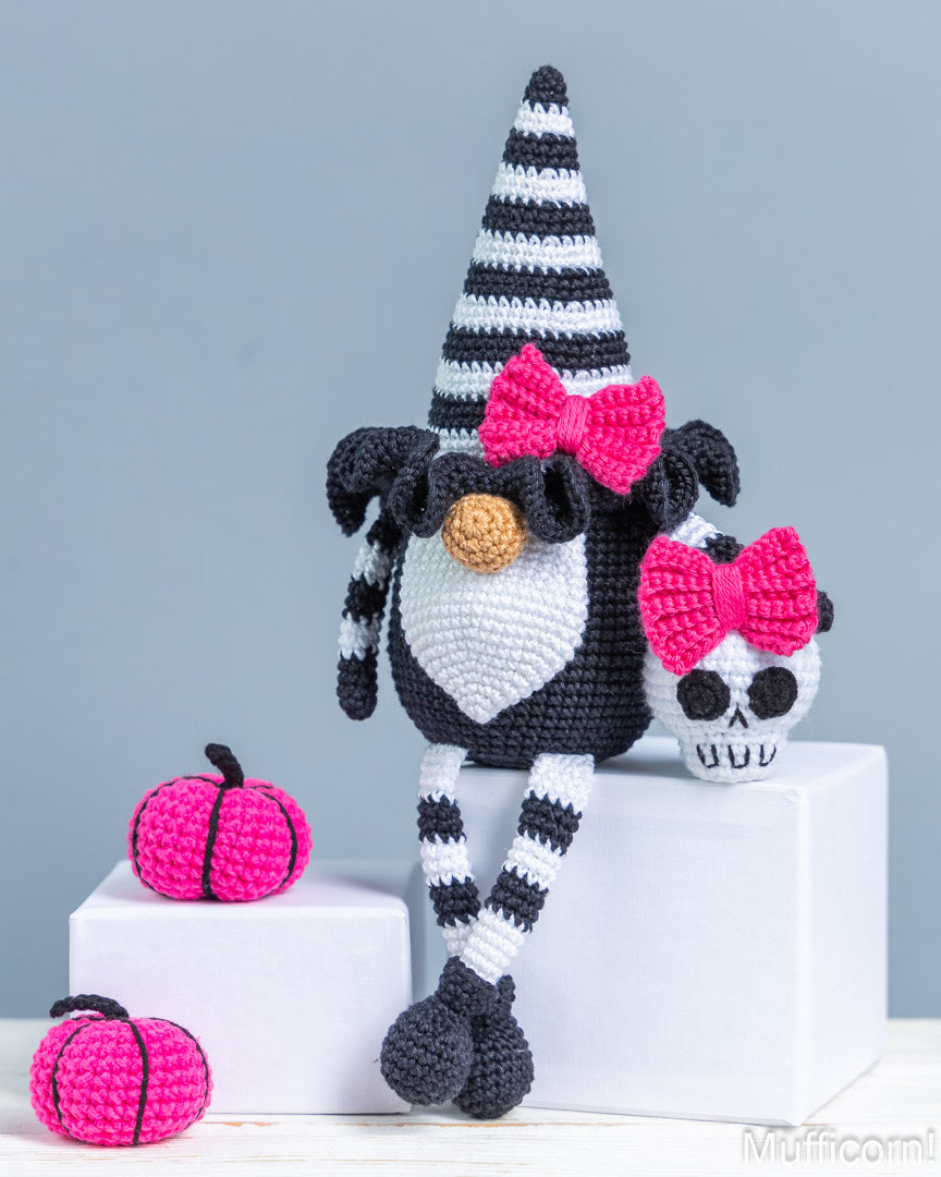 A crocheted Halloween gnome doll with a black-and-white striped hat and legs, pink bow, white face, and small pumpkin companion, displayed on white blocks against a light blue-gray background.