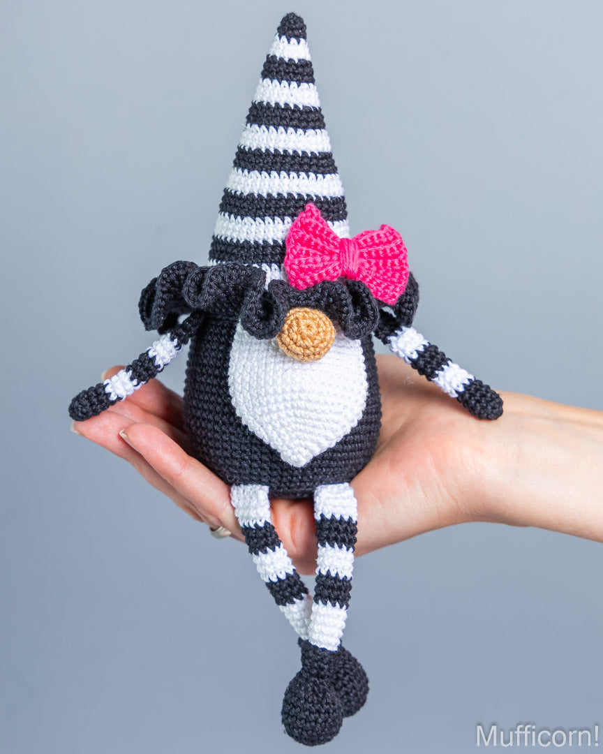 A crocheted gnome doll with a black and white striped hat and legs, a white chest, a bright pink bow, and a round tan nose, being held in a person's hand against a plain gray background.