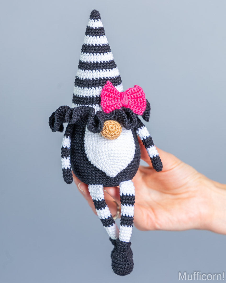 A crocheted gnome doll with a black and white striped hat, body, and striped legs, a white chest, a bright pink bow, and a small tan nose, being held in a person's hand against a plain gray background.