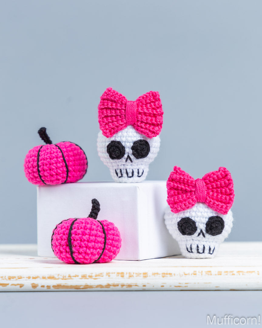 Three crocheted Halloween decorations displayed on a white surface against a light gray background: two bright pink crochet pumpkins with black stems and one white skull wearing a large pink crochet bow. A white display block is used as a prop.