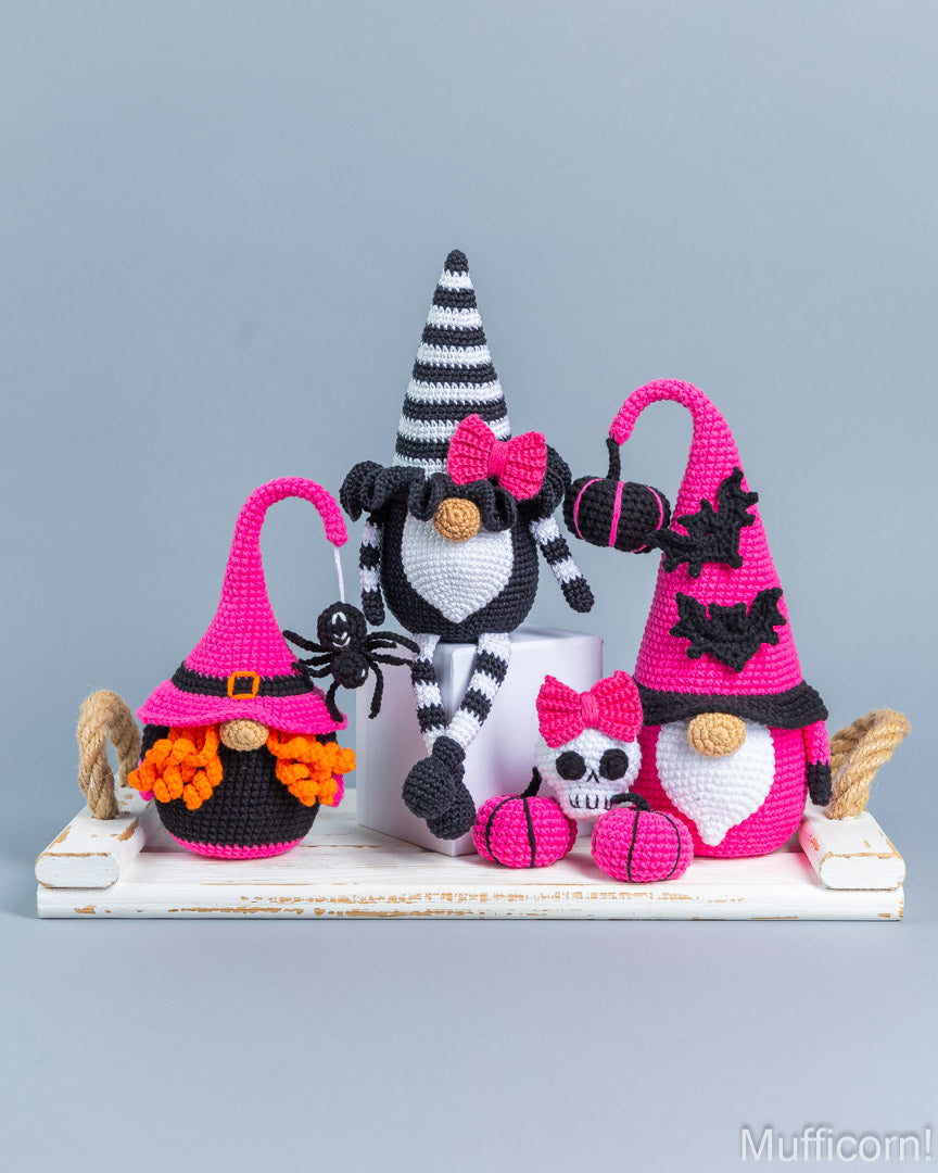 Three crocheted Halloween gnomes displayed on a white wooden tray against a solid gray background. The gnomes feature black and white striped hats, bright pink hats, and are decorated with black bats, orange pumpkins, and small skulls.