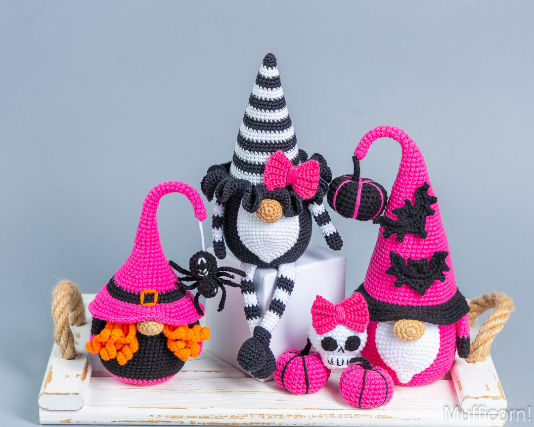 Three crocheted Halloween gnomes displayed on a white wooden tray against a light gray background. The gnomes include a black and white striped gnome with a pink bow, a black and orange gnome in a pink hat, and a pink and white gnome with a tall pink hat and spider accents.