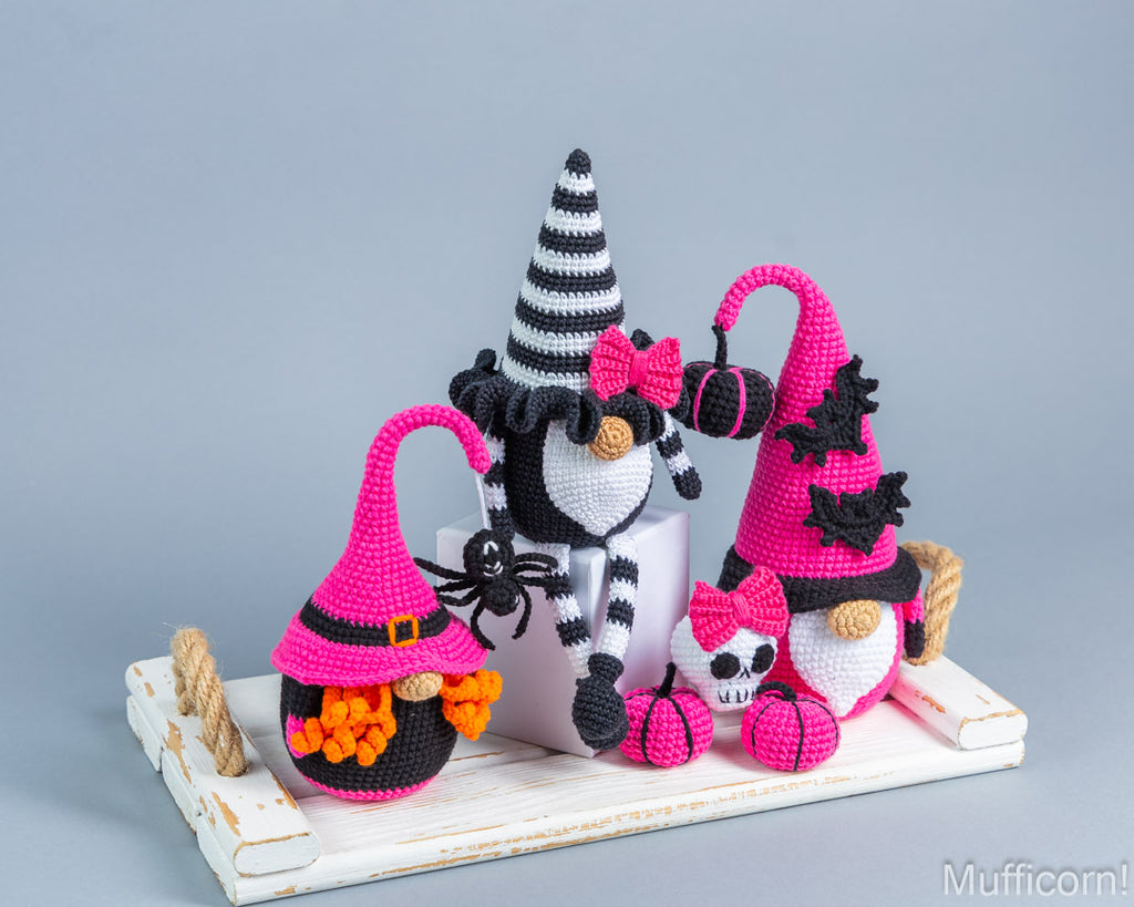 Three crocheted Halloween gnomes  arrangement on a white wooden tray against a light gray background. The gnomes wear black-and-white striped hats, one has a pink bow, one has a pink hat, and one has a skull face.