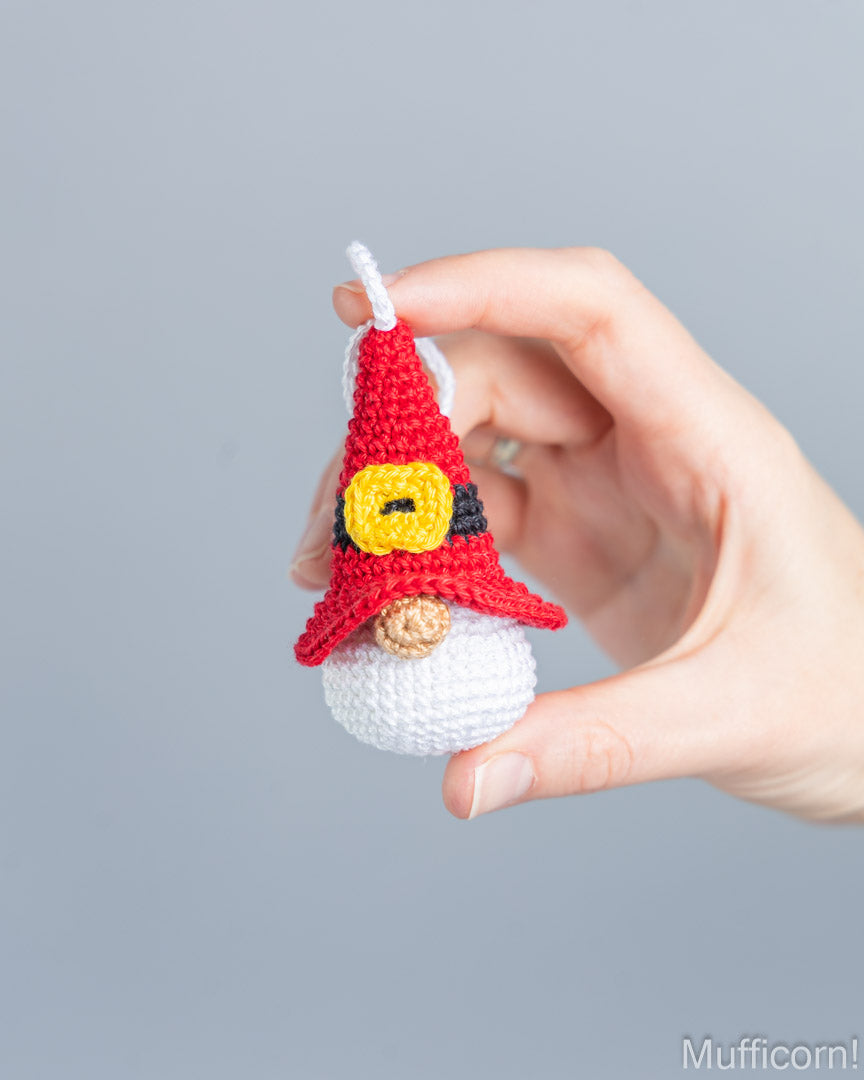 A hand holding a small crocheted santa gnome ornament against a solid gray background. The gnome has a red pointed hat with a yellow and black pattern, a white beard, and a brown nose.