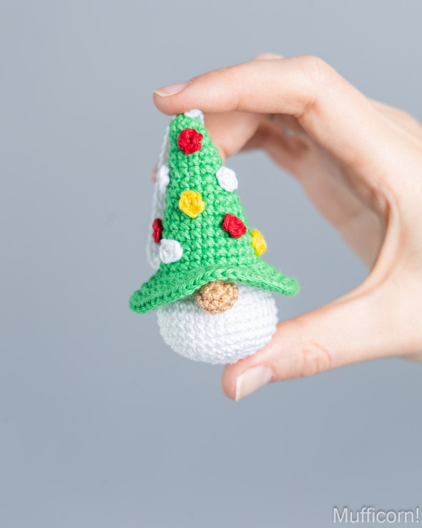 A hand holds a small crocheted gnome wearing a green pointed hat decorated with red, yellow, and white pom-poms, against a plain gray background.