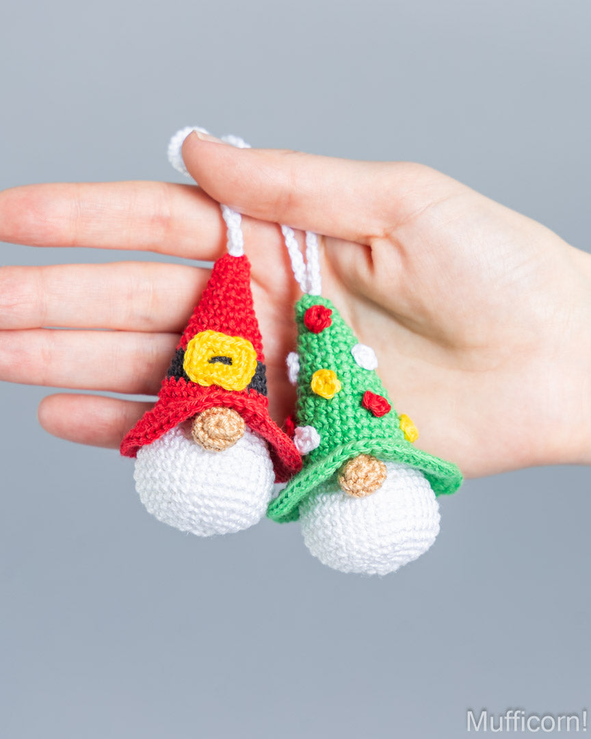 Two small crocheted Christmas gnome ornaments held in a person's hand against a plain gray background. One gnome wears a red hat with a yellow bell detail and white beard, and the other wears a green hat decorated with red, yellow, and white pom-poms and has a white beard.
