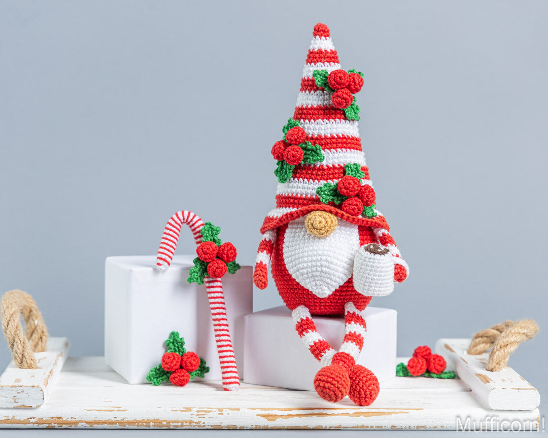 A crocheted Christmas gnome doll with a red-and-white striped hat and legs, green holly accents, and a white beard, posed on white display blocks beside a matching candy cane against a light gray background.