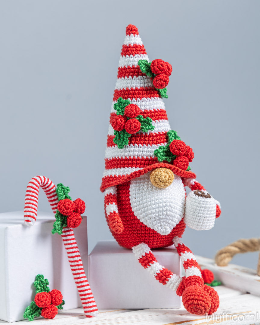 Crocheted Christmas gnome doll with a red-and-white striped hat decorated with green holly leaves and red berries, sitting on a white pedestal. A matching candy cane and small holly sprig rest beside it on a light gray background.