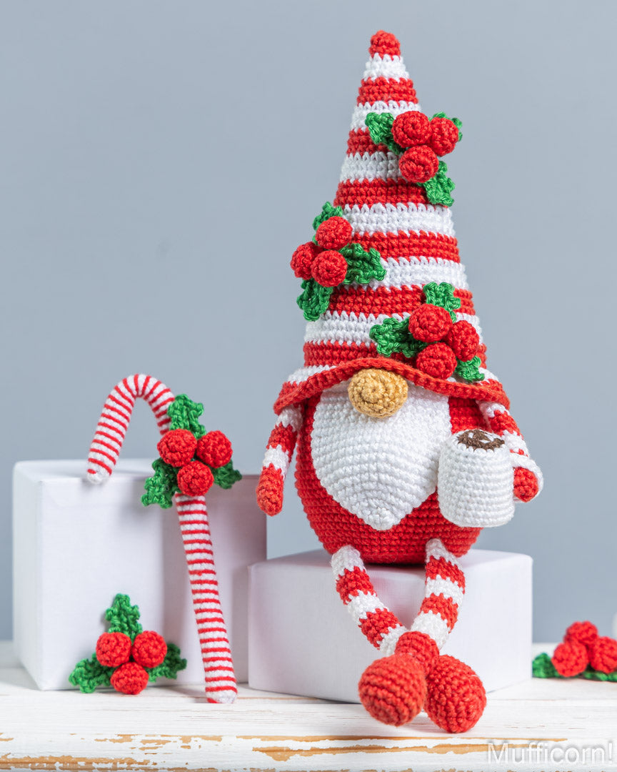 A crocheted Christmas gnome with a red-and-white striped hat, white beard, and red-and-white striped legs, posed on white blocks beside a matching candy cane and small holly decorations against a light gray background.