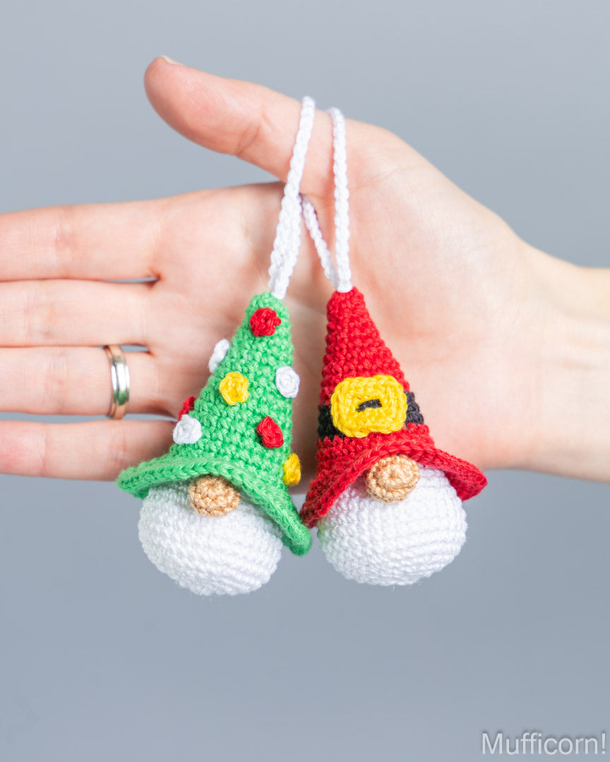 Two crocheted Christmas gnome ornaments held in a person's hand against a plain gray background. One gnome wears a green hat with yellow and red ornaments, and the other wears a red hat with a yellow belt detail.