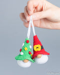 A close-up of a hand holding two small crocheted Christmas gnome ornaments against a plain gray background. One gnome is shaped like a green Christmas tree with red, yellow, and white accents, and the other is a red gnome with a yellow hat and white beard.