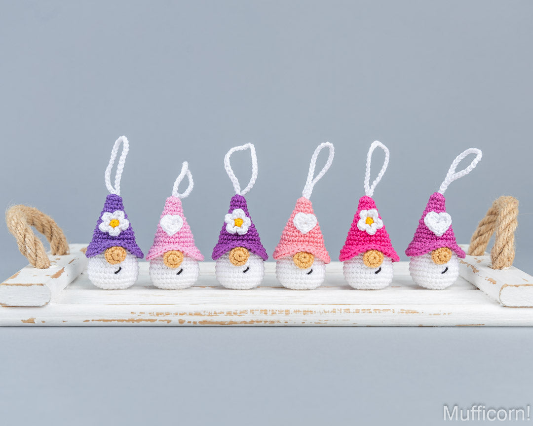 Six small crocheted gnome ornaments in purple and pink tones are lined up on a white wooden tray with rope handles, against a light gray background. The Mufficorn logo appears in the bottom right corner.