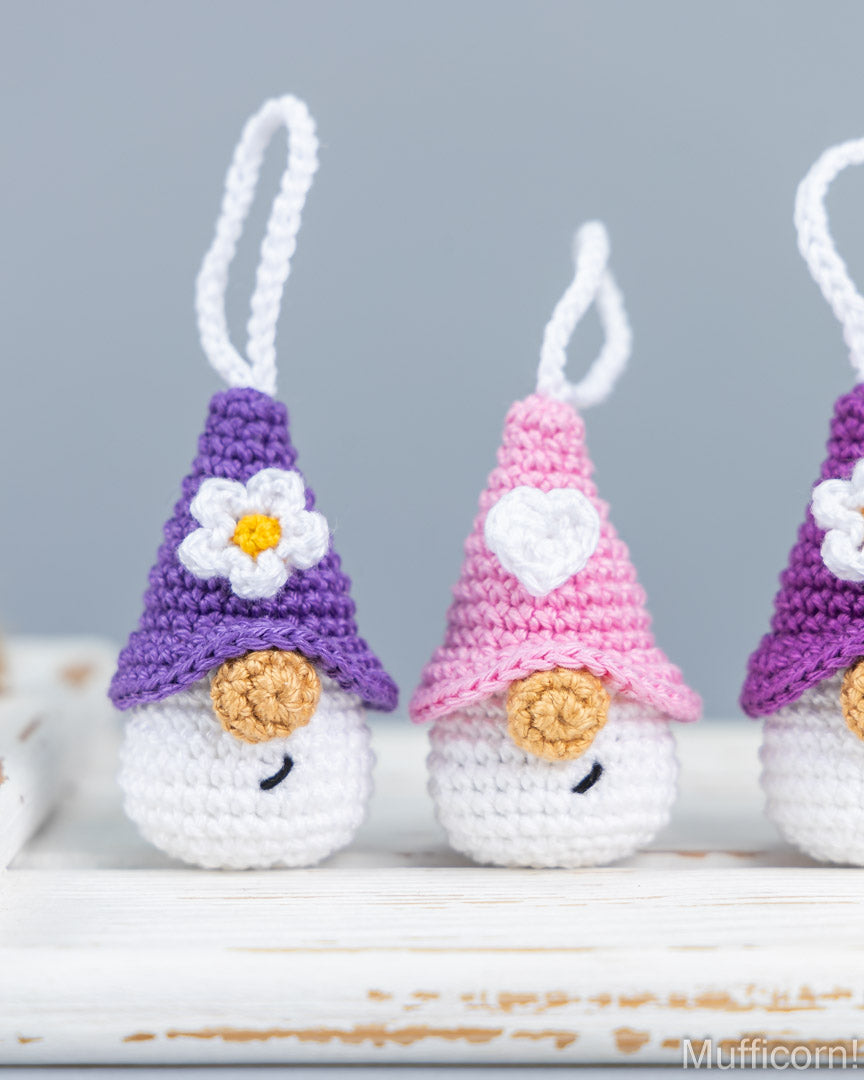 Three small crocheted gnome ornaments with white hanging loops are lined up on a white rustic surface against a soft gray background. The center gnome wears a pink hat with a white heart, and the side gnomes wear purple hats with white flowers.