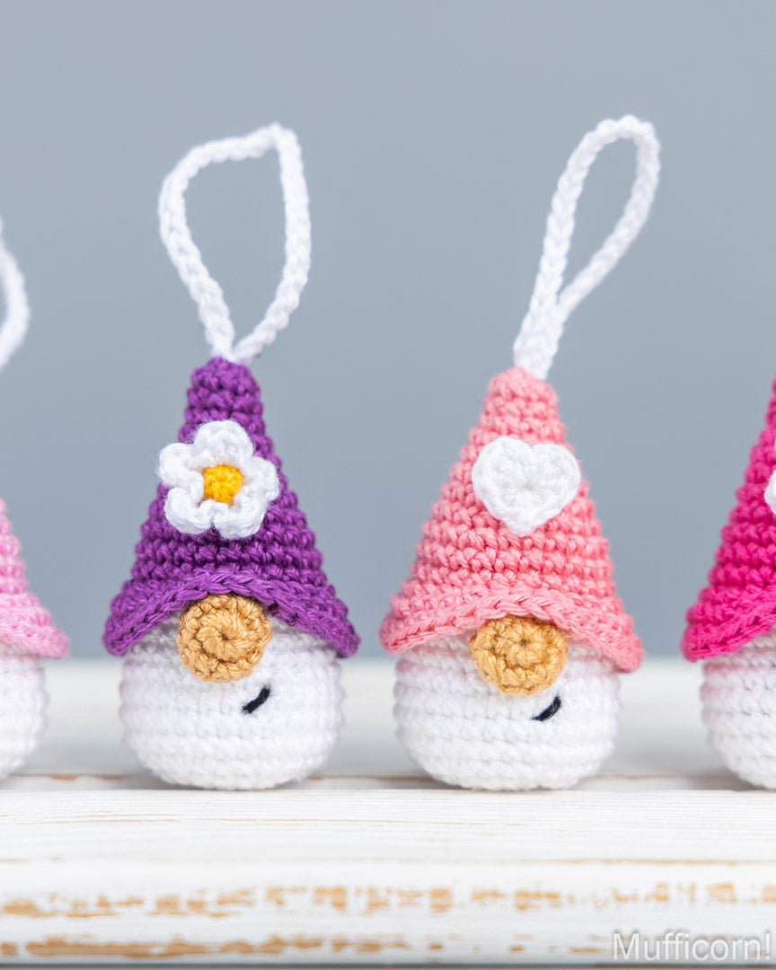 Three small crocheted gnome ornaments with white hanging loops, shown in purple, pink, and magenta on a white wooden surface against a soft gray background.
