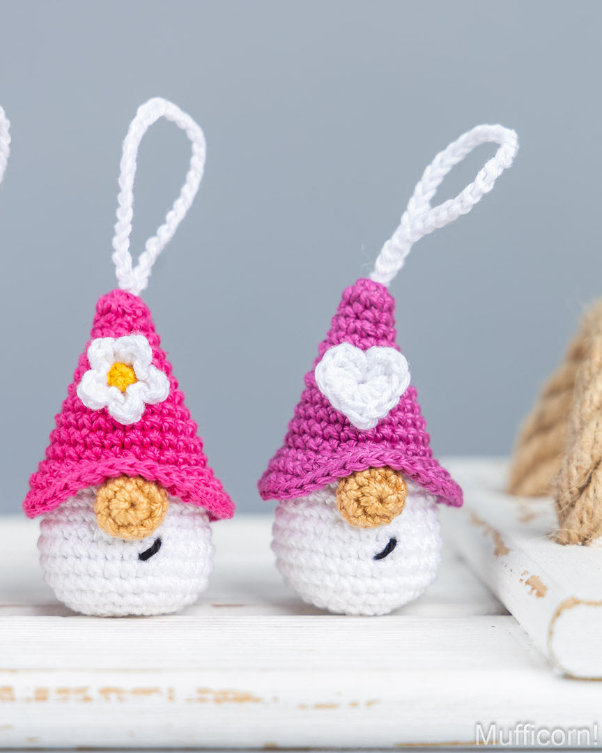 Two small crocheted gnome ornaments with white loops at the top, wearing pink and purple pointed hats decorated with white flowers and hearts, standing on a white wooden surface against a soft gray background.