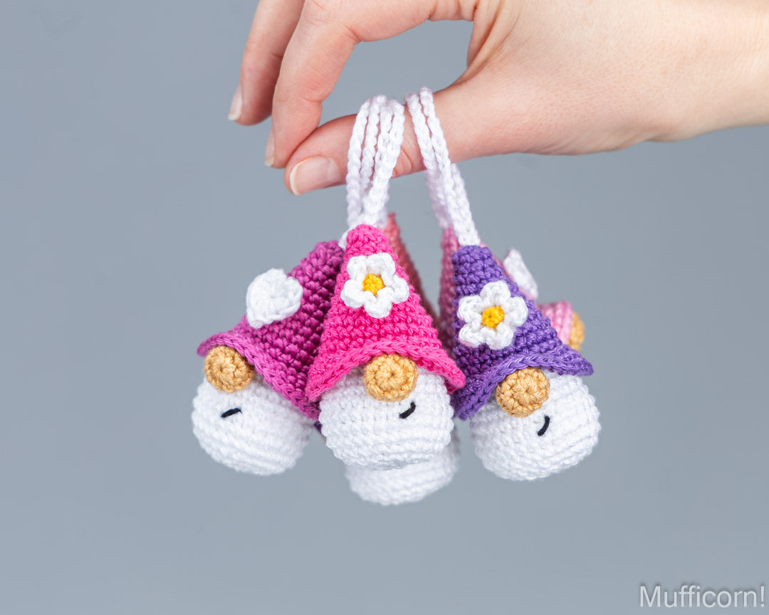 Three small crocheted mufficorn character keychains held by a hand against a solid gray background. The keychains have white bodies, pink and purple hats, white daisy flower details, and small yellow buttons.