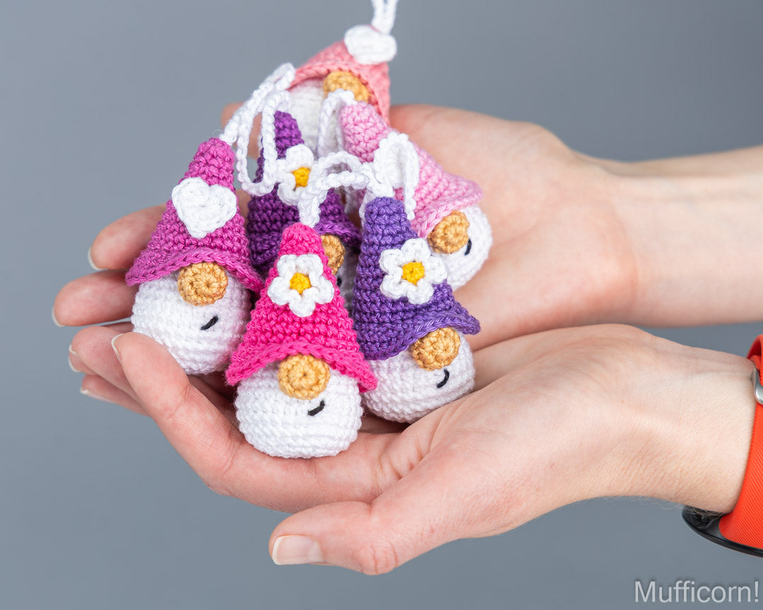 A pair of hands holding a cluster of small crocheted gnome ornaments in pink, purple, and white, each with a flower on its hat, against a plain gray background.