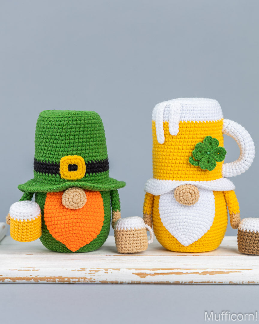 Two crocheted St. Patrick's Day-themed mug covers shaped like a leprechaun and a beer mug, displayed on a rustic white wooden surface against a solid gray background.
