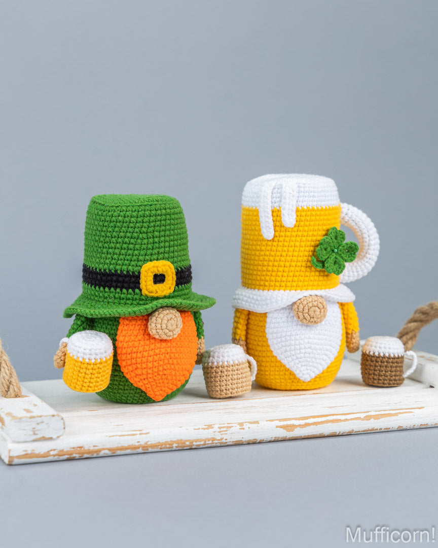 Two crocheted St. Patrick's Day gnome mug covers on a distressed white wooden surface against a solid gray background.
