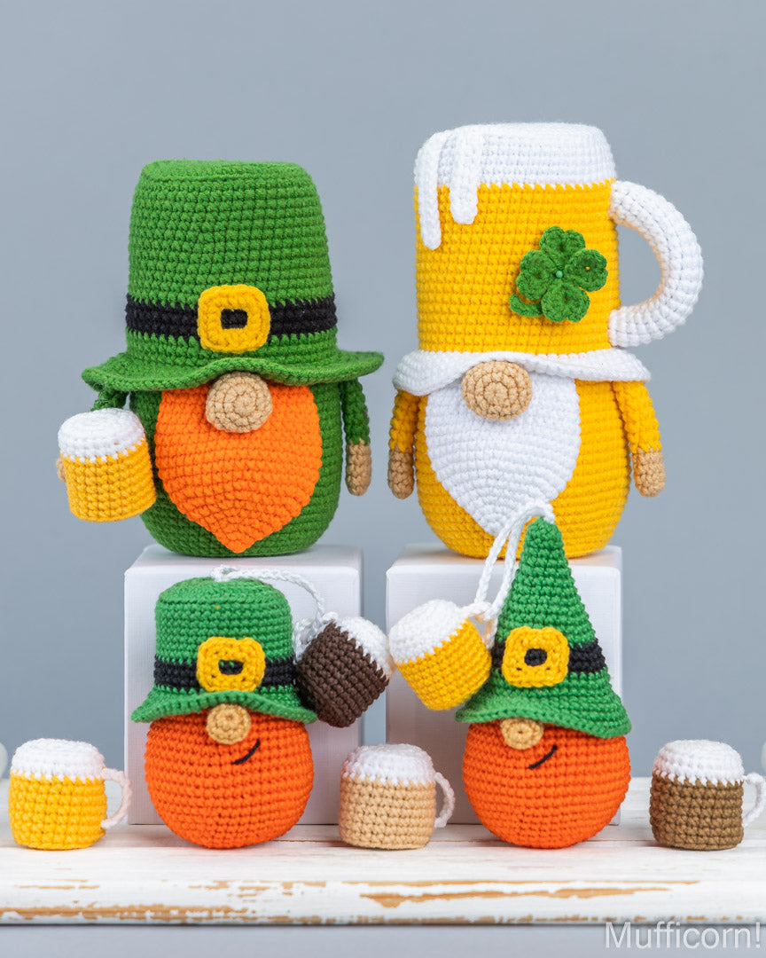 Four crocheted St. Patrick's Day leprechaun mug cozies and beer mug accessories arranged on white blocks against a light gray background.