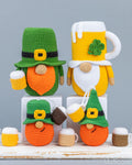 Four crocheted St. Patrick's Day leprechaun mug cozies and beer mug accessories arranged on white blocks against a light gray background.