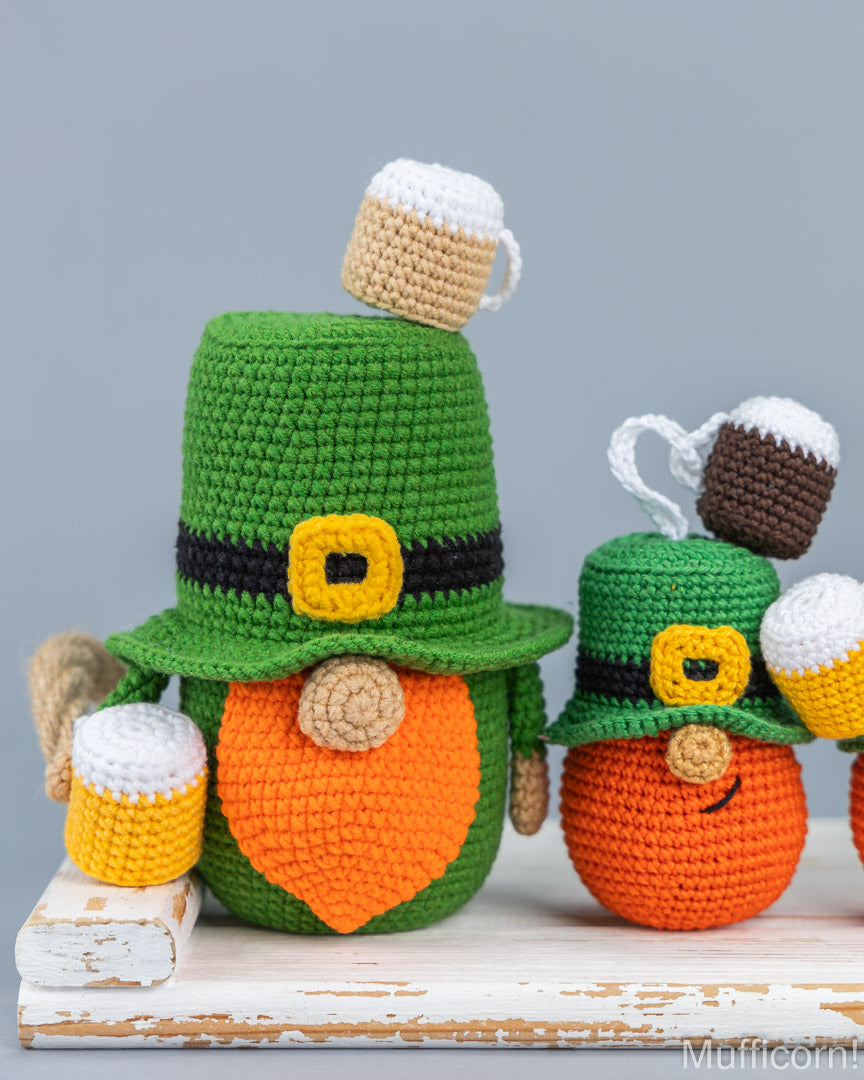 Two crocheted leprechaud-style figures with green hats, orange faces, and beer mug accessories, displayed on a rustic white wooden surface against a light gray background.