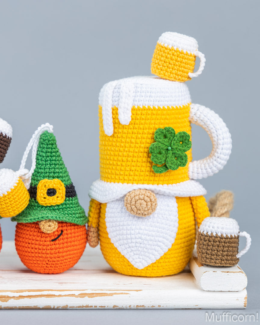 A crocheted St. Patrick's Day themed beer mug gnome cover sits on a white wooden surface next to a small orange gnome and a matching miniature mug, against a solid gray background.