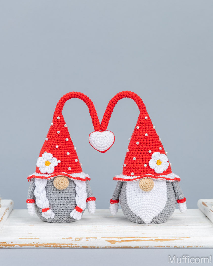 Two crocheted gnome figures with red pointed hats, white beards, gray bodies, and white flower accents, posed on a distressed white wooden surface against a light gray background.