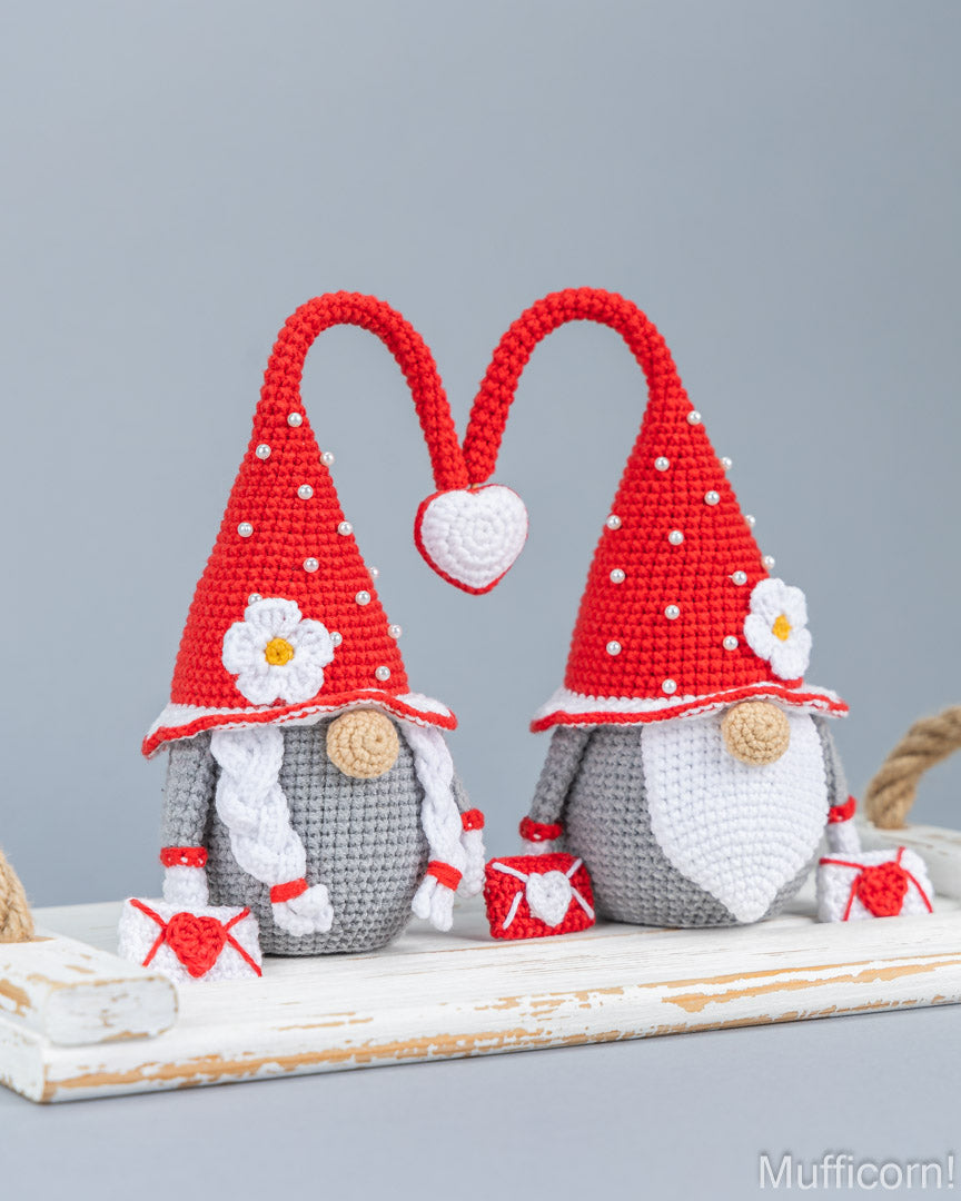 Two crocheted gnomes with red pointed hats, white beards, gray bodies, and white flowers, posed together on a rustic white wooden surface against a light gray background.