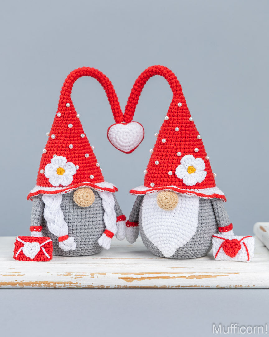 Two crocheted gnomes with tall red hats and white beards, posed together on a rustic white surface against a light gray background. Each gnome has a red hat with white pom-poms, a white flower detail, and a small red-and-white gift bag.