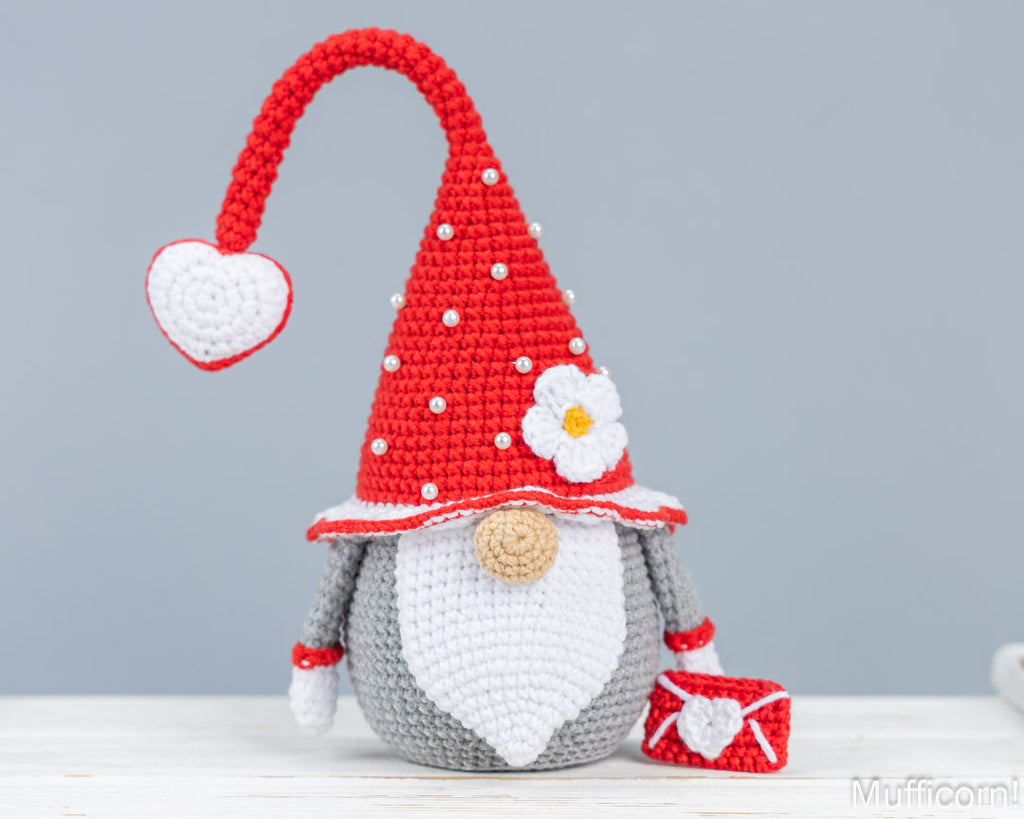A crocheted gnome doll with a red pointed hat, white beard, gray body, and small red-and-white envelope, photographed on a light gray background.