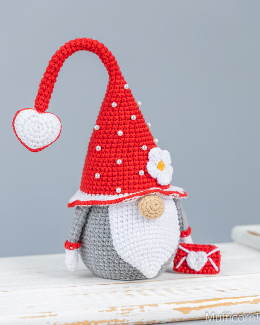 A crocheted gnome figurine with a red pointed hat, white beard, gray body, and a small red-and-white heart-shaped accessory, displayed on a white wooden surface against a soft gray background.