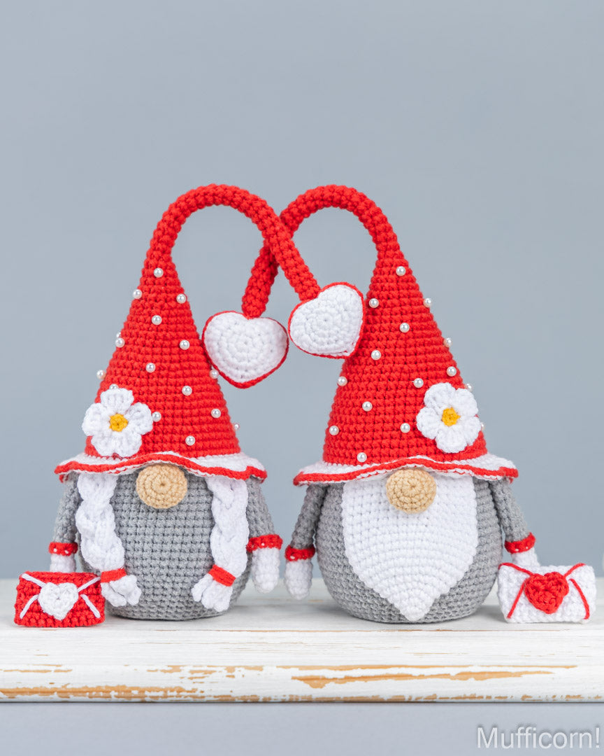 Two crocheted gnome figures with tall red pointed hats, white beards, white flowers, and small red-and-white accessories, displayed on a distressed white surface against a light gray background.