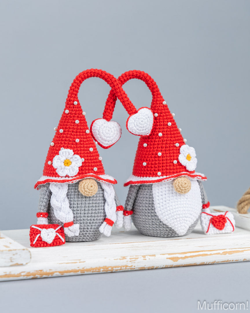 Two crocheted gnome figures with red pointed hats, white beards, and small red-and-white accessories, displayed on a rustic white wooden surface against a soft gray background.