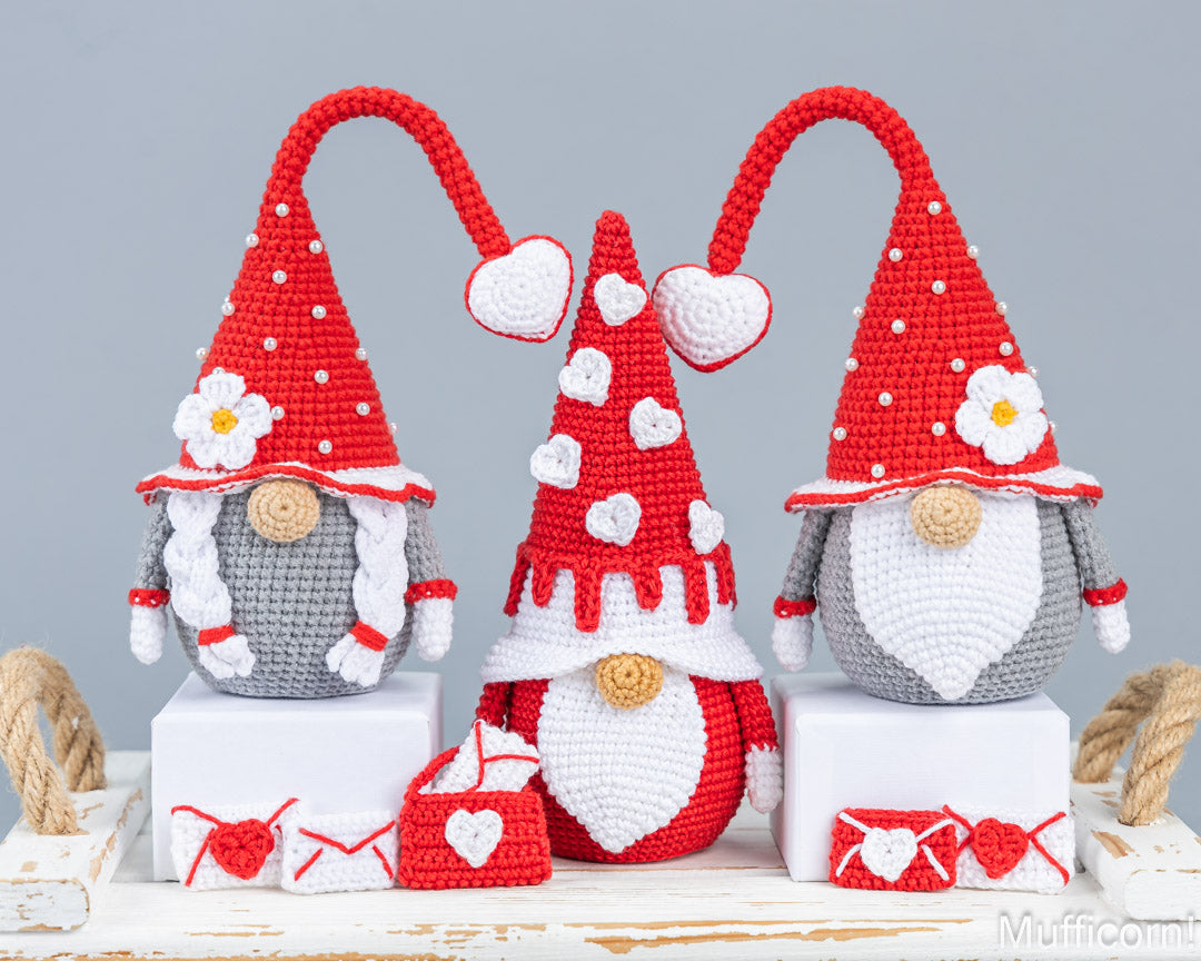Three crocheted gnome figurines in red, white, and gray are displayed on white blocks with small red and white heart-themed accessories in front, set against a plain gray background.