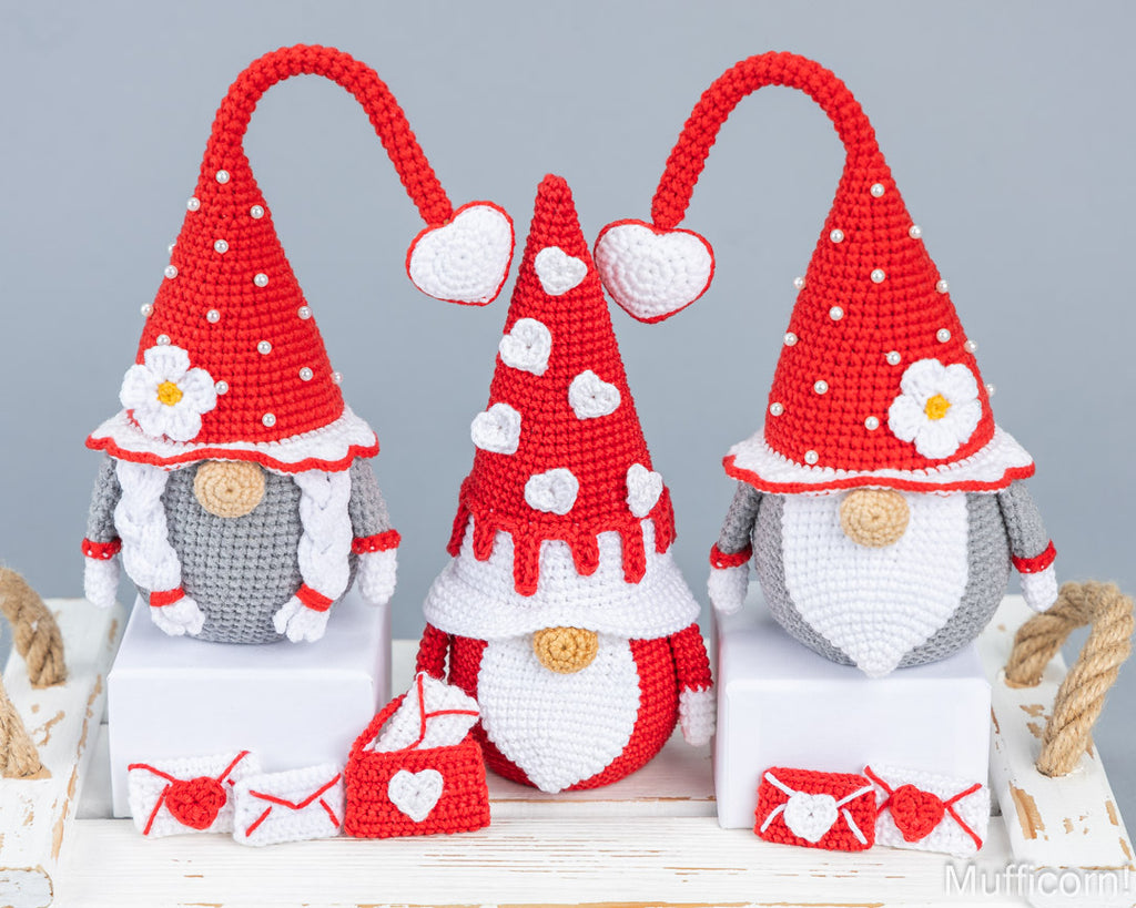 Three crocheted Valentine's Day gnome ornaments displayed on white blocks, featuring red and white hats with hearts and flowers, gray bodies, and small red-and-white accessories on a rustic white table against a light gray background.