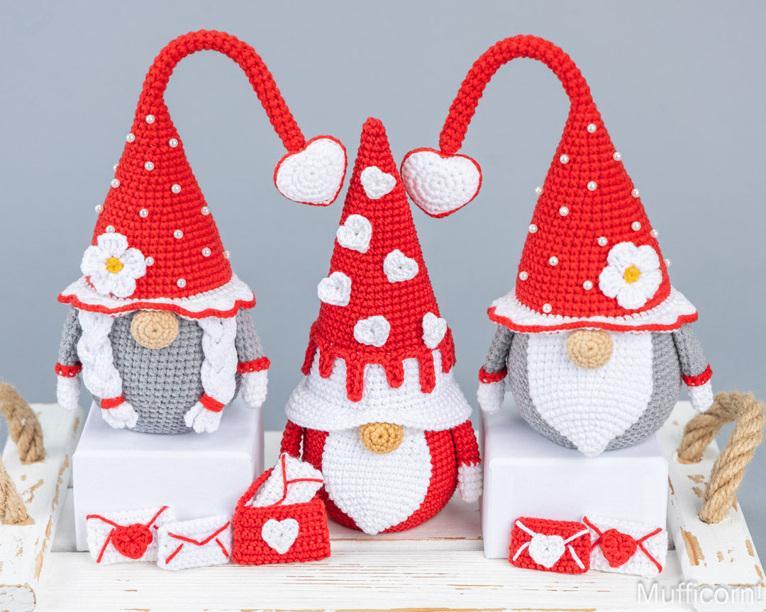 Three crocheted Valentine's Day gnome ornaments displayed on white blocks, featuring red and white hats with hearts and flowers, gray bodies, and small red-and-white accessories on a rustic white table against a light gray background.