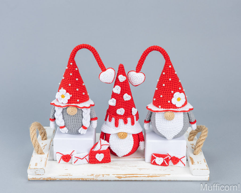 Three crocheted Christmas gnomes in red and white with gray beards, displayed on a white rustic wooden tray against a solid gray background.