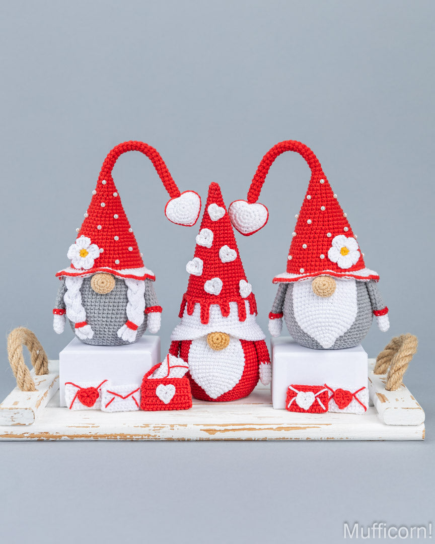 Three crocheted Christmas gnomes in red, white, and gray, arranged on a white rustic tray with small red and white heart-themed accessories against a solid gray background.