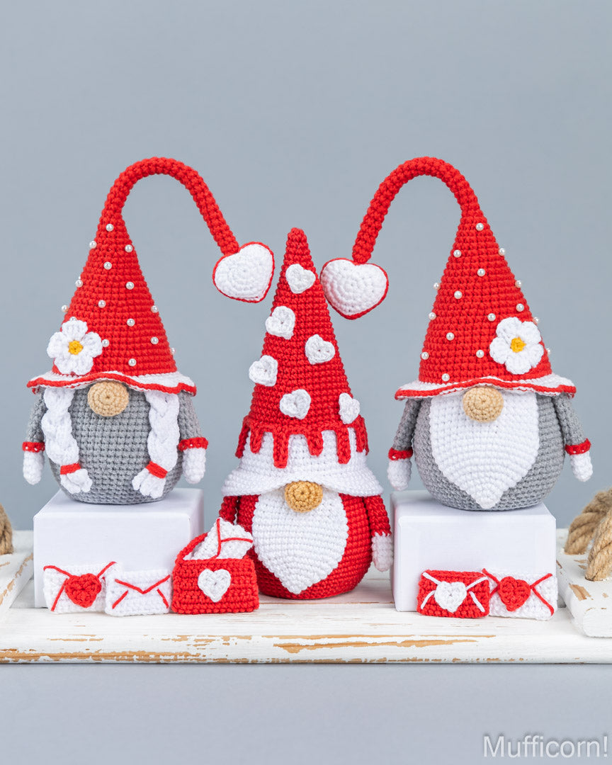 Three crocheted gnome decorations in red, white, and gray are displayed on white pedestals against a light gray background. The gnomes have tall pointed hats with white dots, white beards, and small heart and flower details, with matching red and white heart-themed accessories in front.