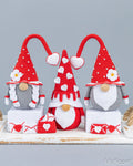 Three crocheted gnome decorations in red, white, and gray are displayed on white pedestals against a light gray background. The gnomes have tall pointed hats with white dots, white beards, and small heart and flower details, with matching red and white heart-themed accessories in front.