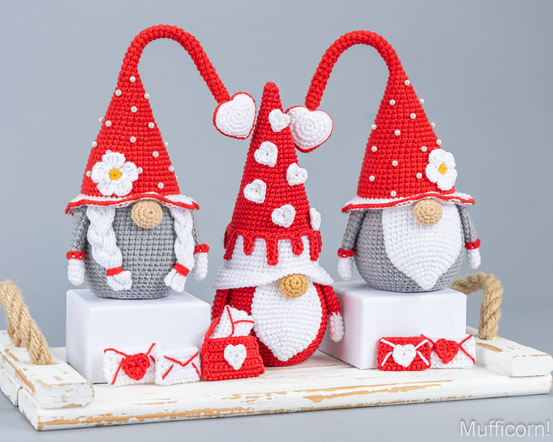 Three crocheted gnome decorations arranged on a white wooden tray against a light gray background. The gnomes are red and white with gray bodies, white beards, and small white flowers on their hats.
