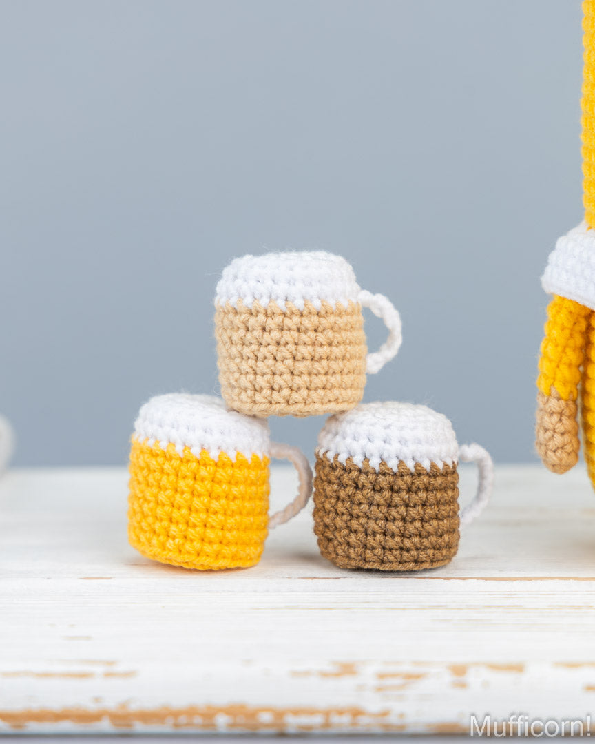 Three crocheted beer mug toys stacked on a white wooden surface against a soft gray background. The mugs are colored in yellow, brown, and tan, with white foam tops.