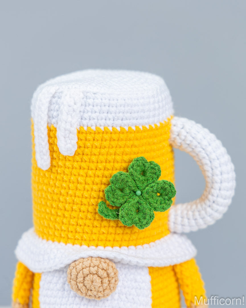A close-up of a crocheted beer mug character, likely a St. Patrick's Day decoration or plush toy, featuring a yellow body, white frothy top, a green four-leaf clover, and a beige button detail, set against a solid gray background.